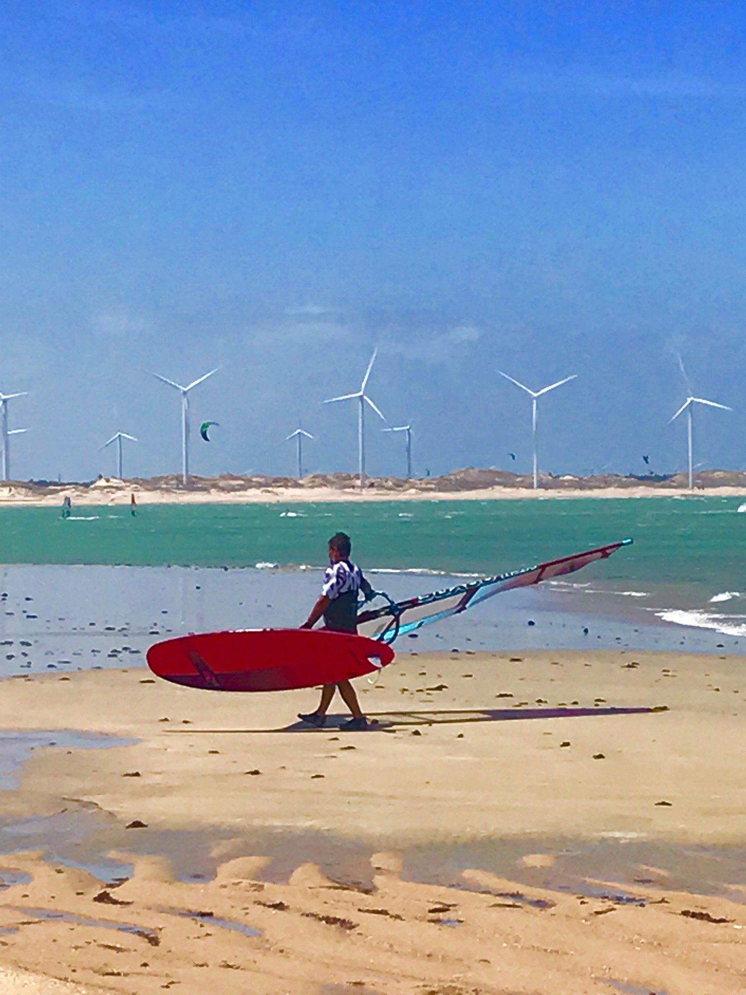 Um homem carregando uma prancha de surfe vermelha em uma praia com moinhos de vento ao fundo
