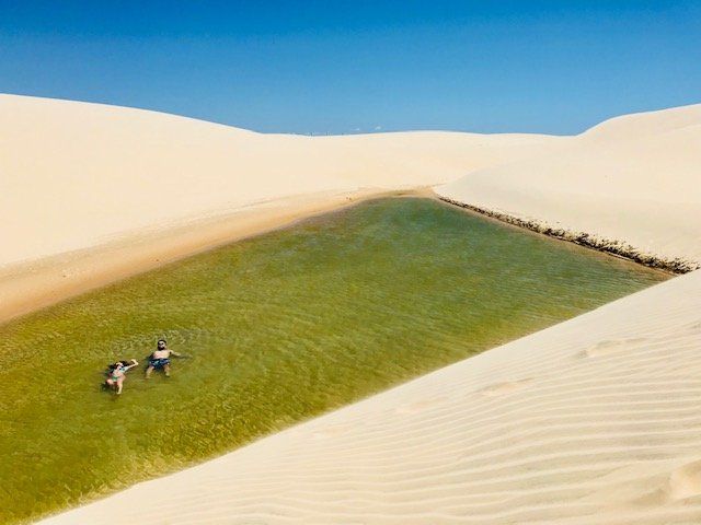 Algumas pessoas em pé no topo de uma duna de areia ao lado de uma poça de água.
