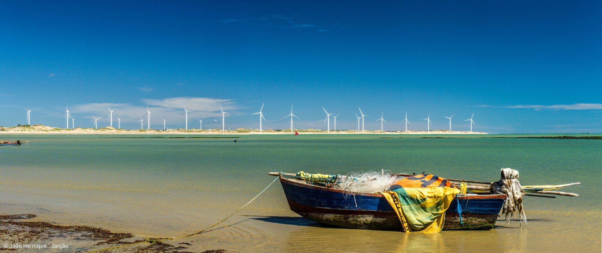 Um barco está amarrado à margem de um corpo d'água com turbinas eólicas ao fundo.