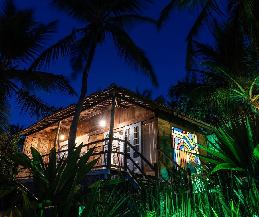 Uma cabana rústica de madeira situada em meio a altas palmeiras e vegetação tropical exuberante, em uma praia de areia sob um céu azul brilhante.