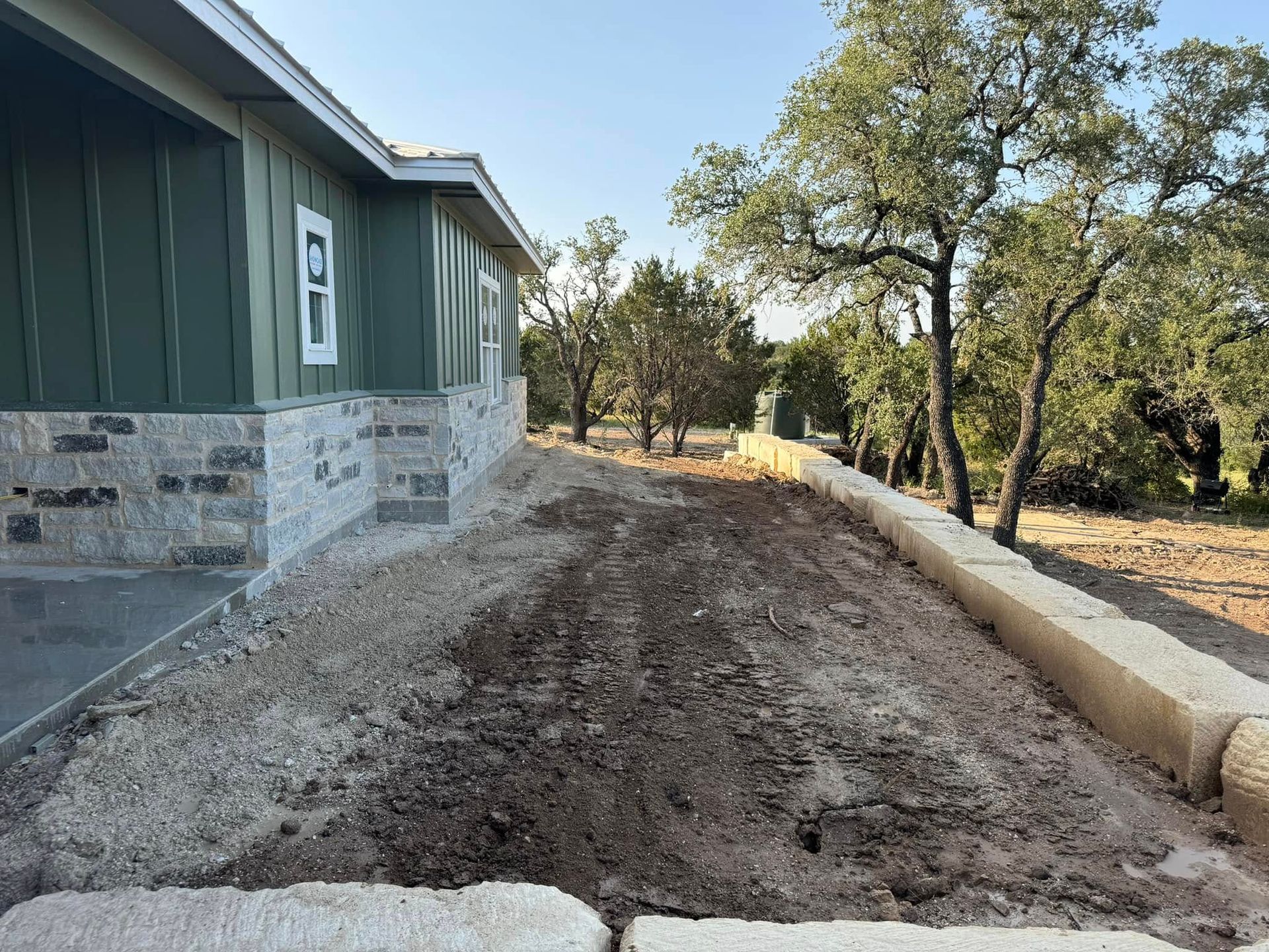 Exterior of a green building with stone accents. A dirt path runs along the side, bordered by a stone wall, trees in background.