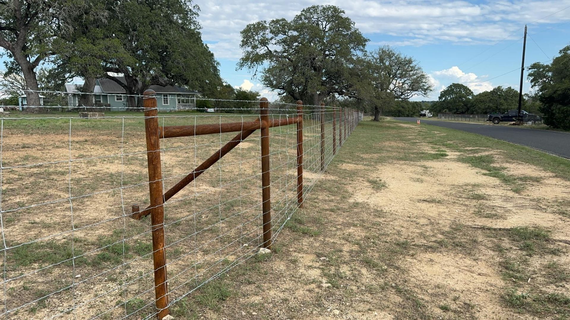 Rusty metal fence along a dirt road with a house and trees in the background.