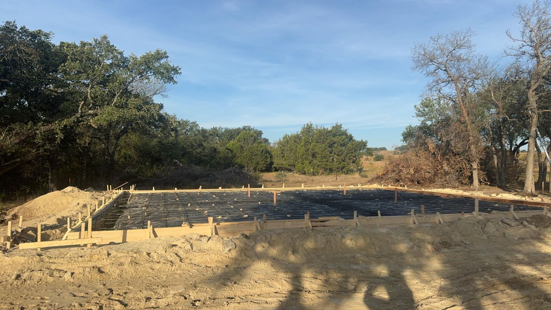 Construction site with wooden forms and black fabric, surrounded by dirt and trees under a blue sky.