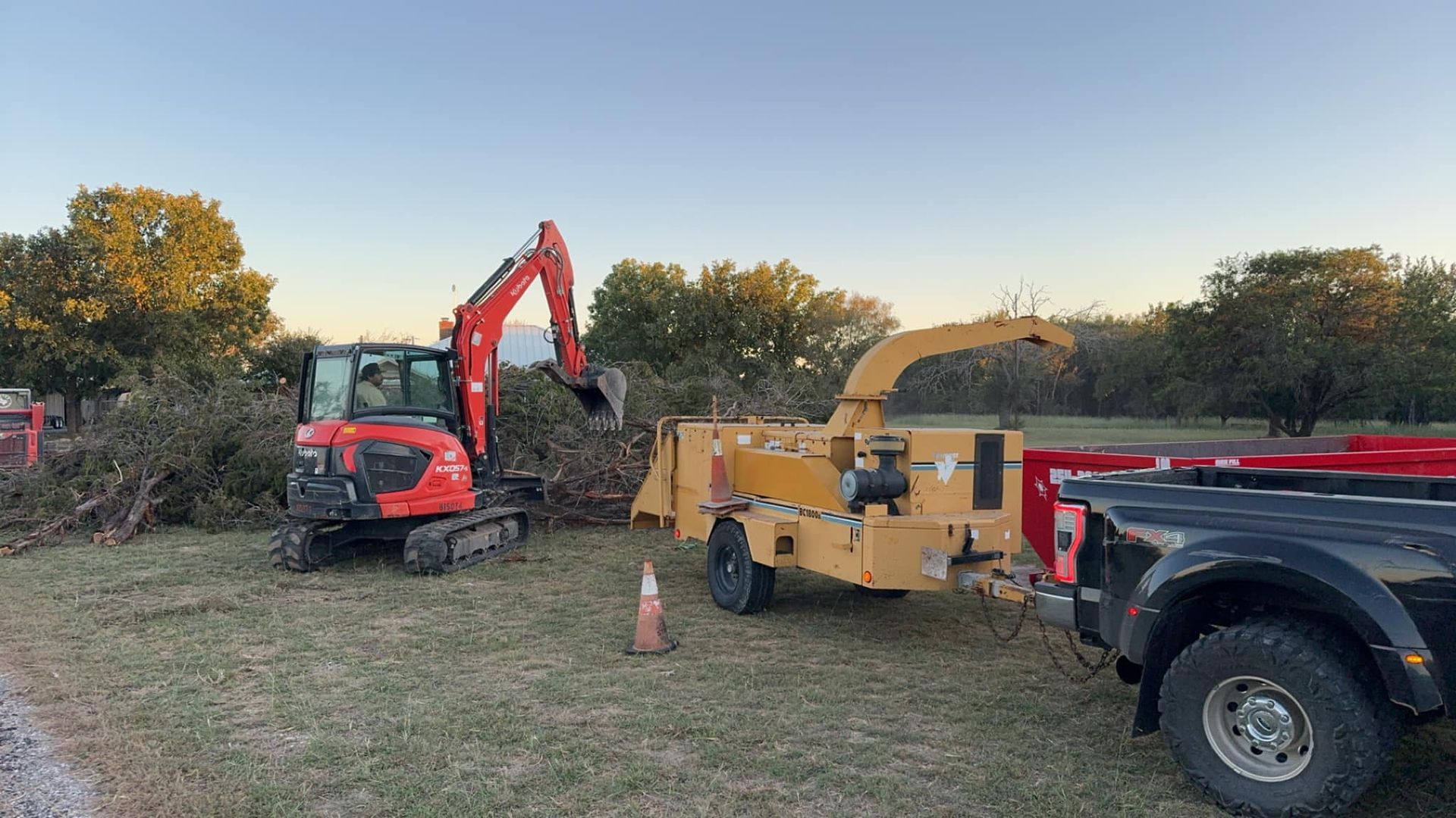A red excavator loading tree branches into a wood chipper pulled by a black truck on a grassy field.