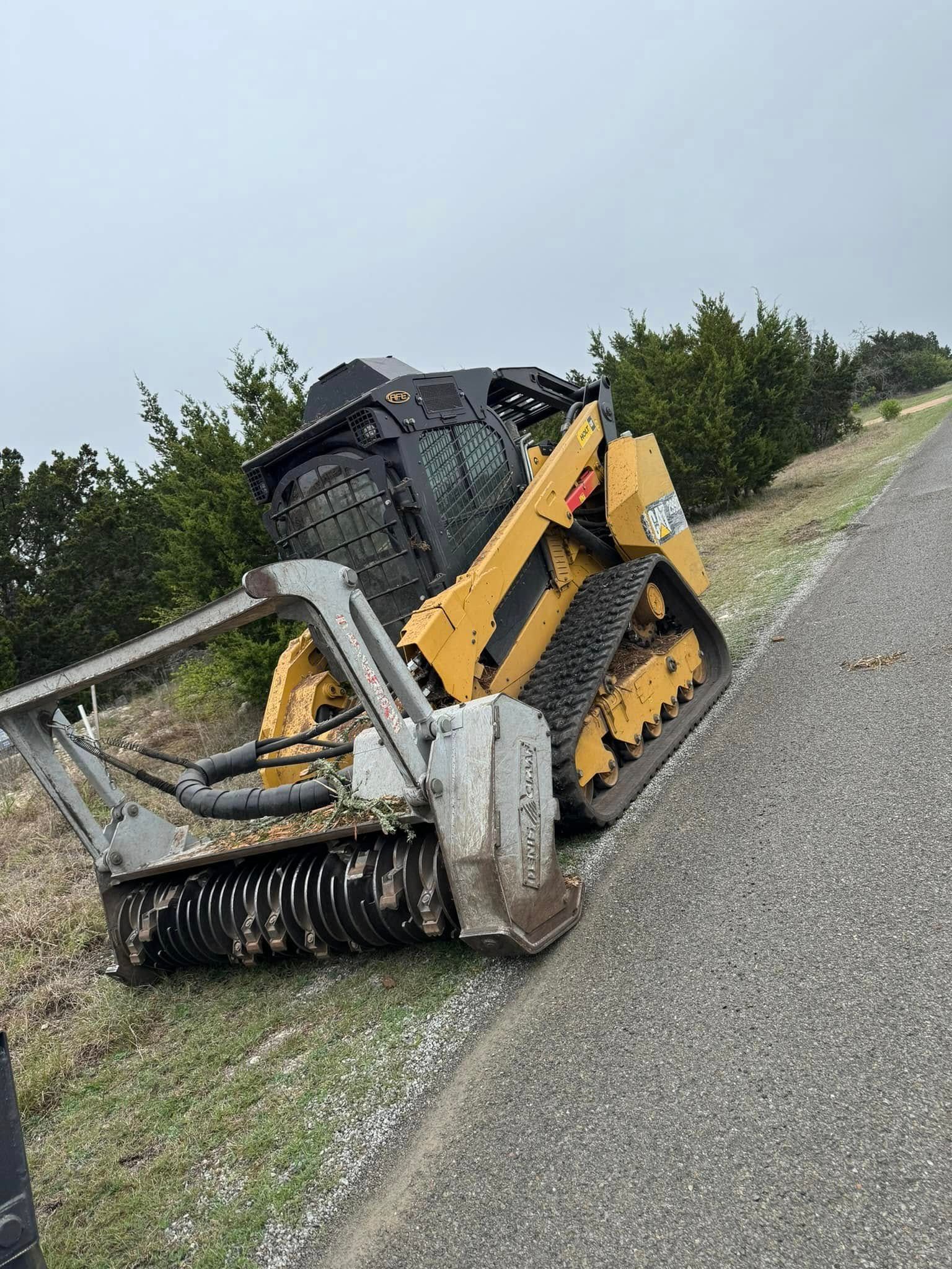 Yellow skid steer with brush cutter attachment on a roadside, tilted on an incline.
