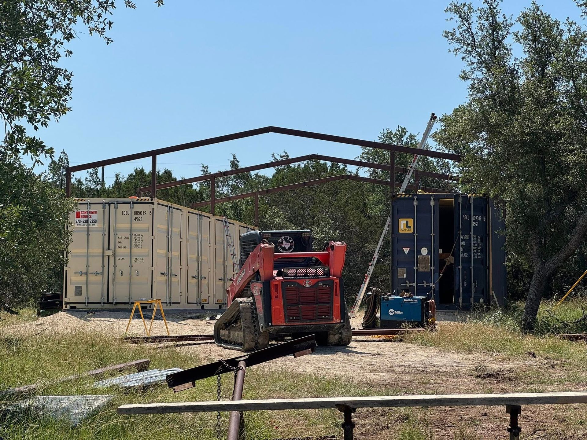 Red Bobcat in front of shipping containers under metal frame; rural outdoor setting.