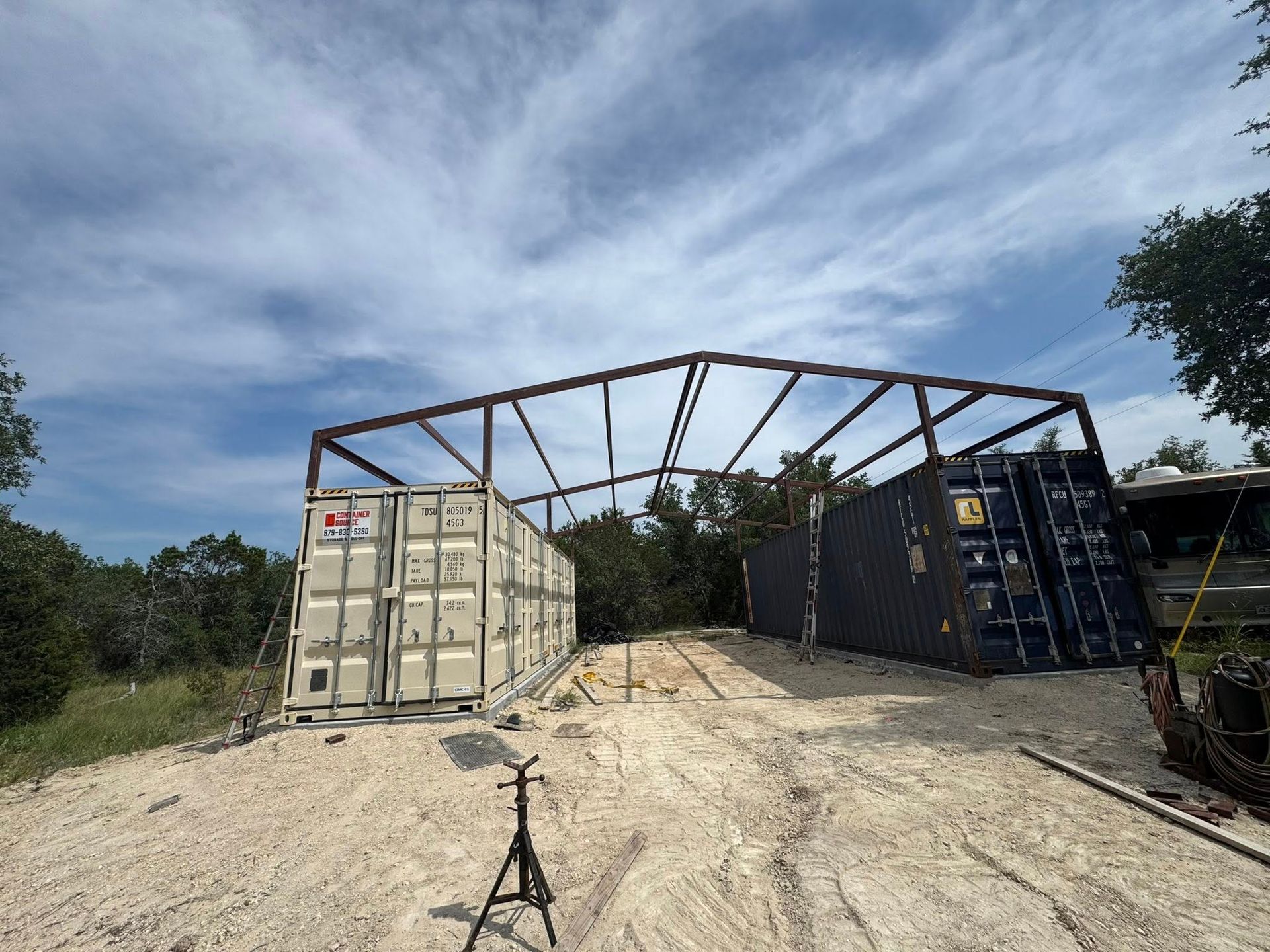 Two shipping containers with a metal frame roof on a dirt lot under a cloudy sky.