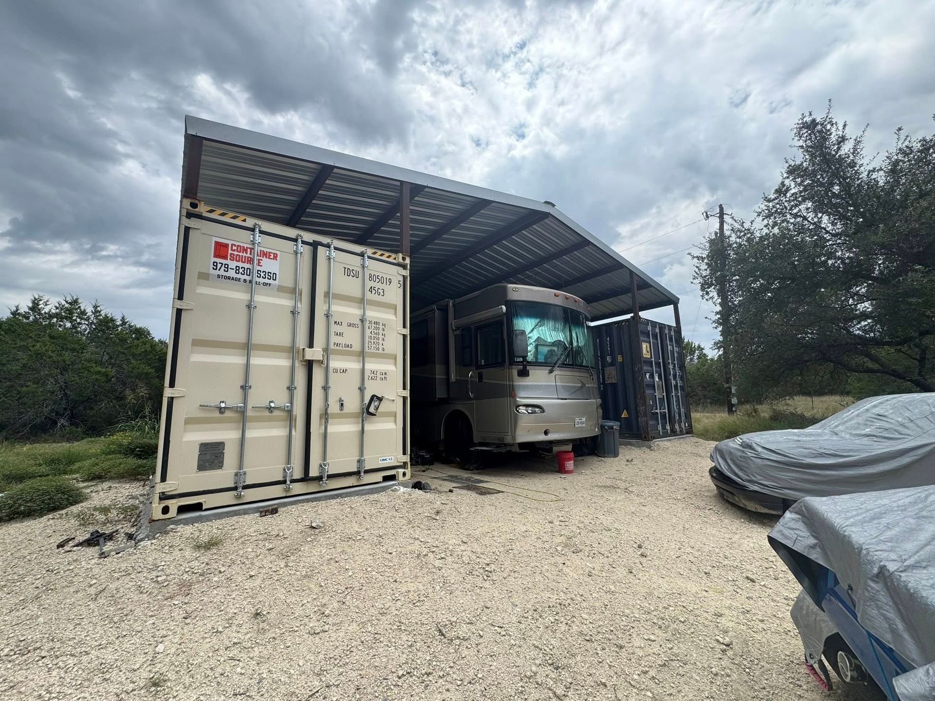 RV parked under a metal roof shelter next to shipping containers on a gravel lot. Cloudy sky.