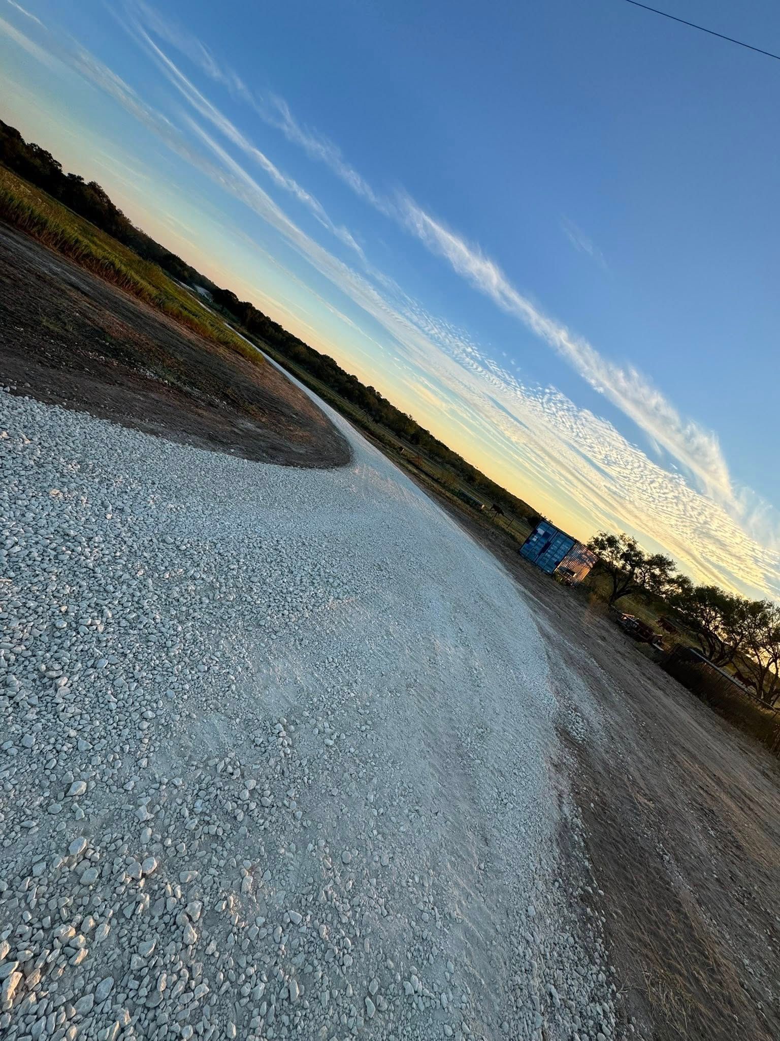 Gravel road curves into the distance beneath a blue sky with streaky clouds; trees and fields line the edges.
