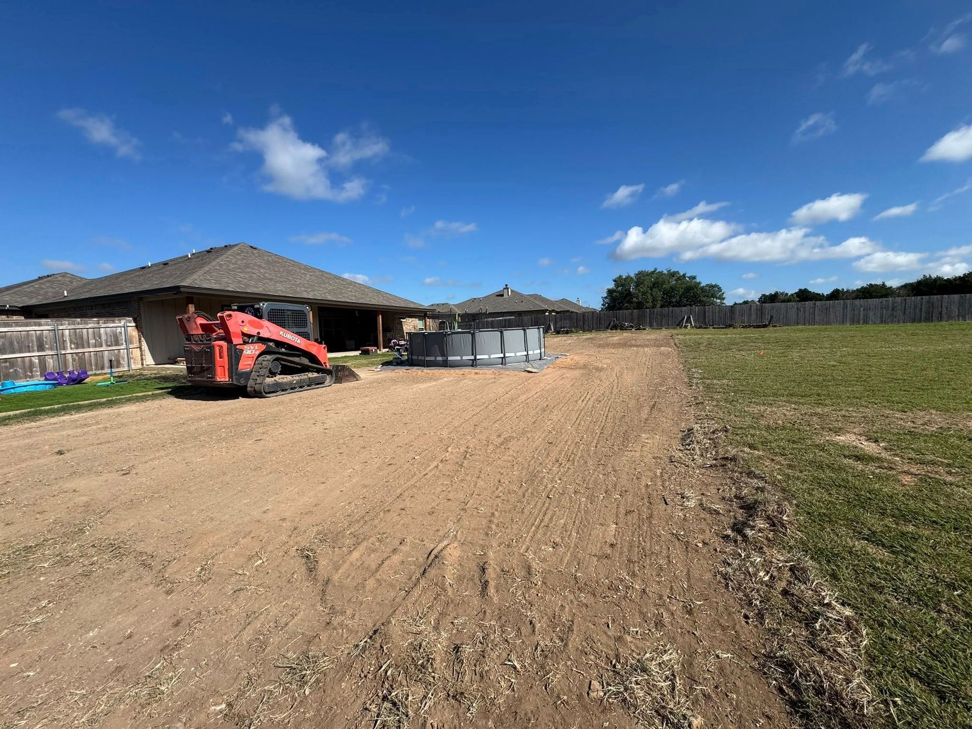 A construction site with an orange skid steer, dirt path, and a partially constructed above-ground pool under a blue sky.