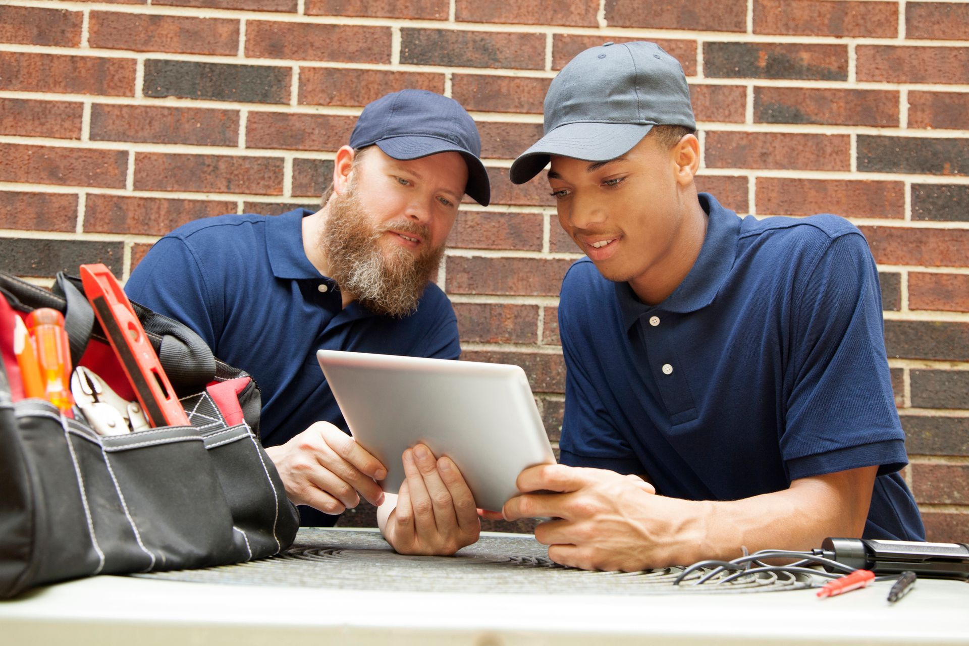 Two technicians standing over a home's air conditioner unit outdoors and looking at a digital tablet