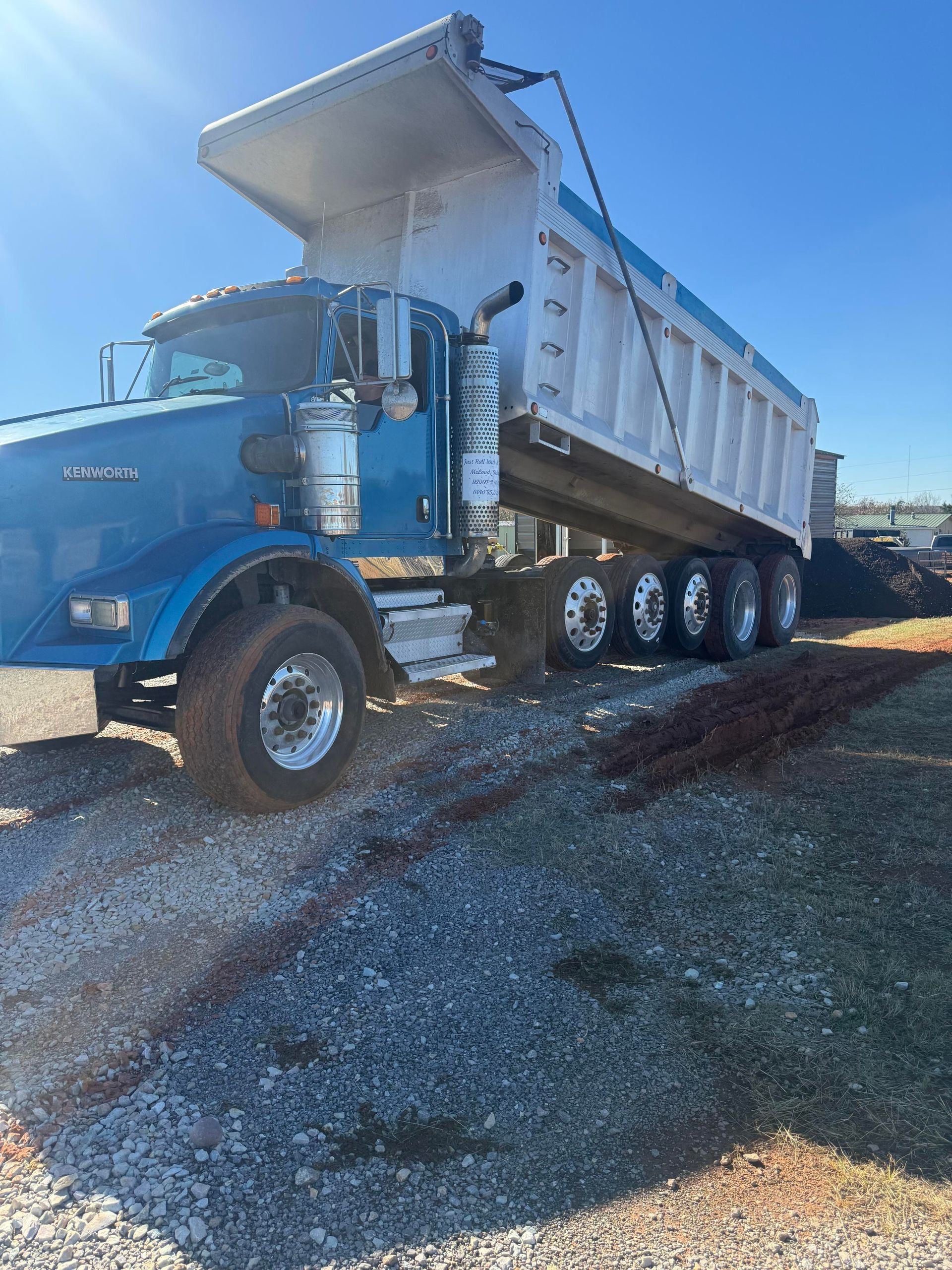 Construction site: Dump truck with driver, worker holding cable, another truck in background. Dusty, sunny day.