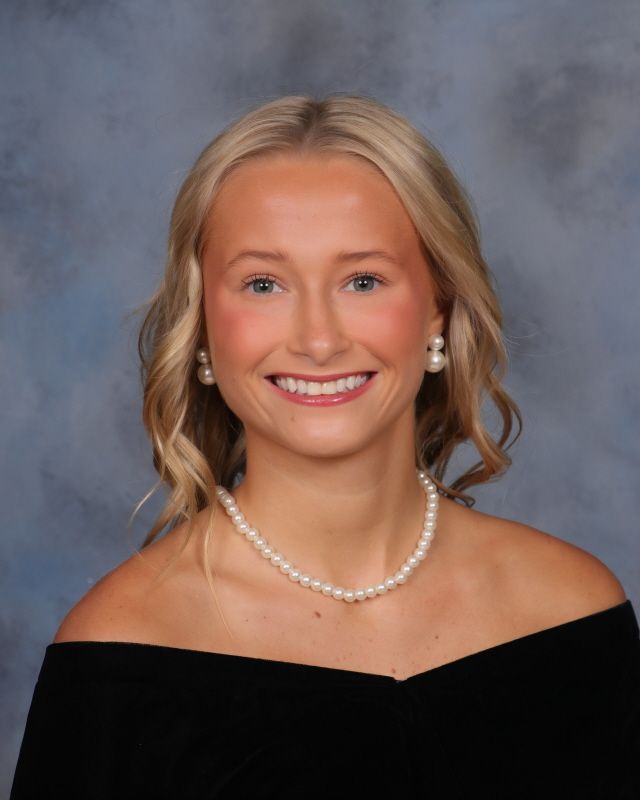 Headshot of a person with wavy blonde hair, wearing a black off-the-shoulder top and pearl necklace, smiling at camera.