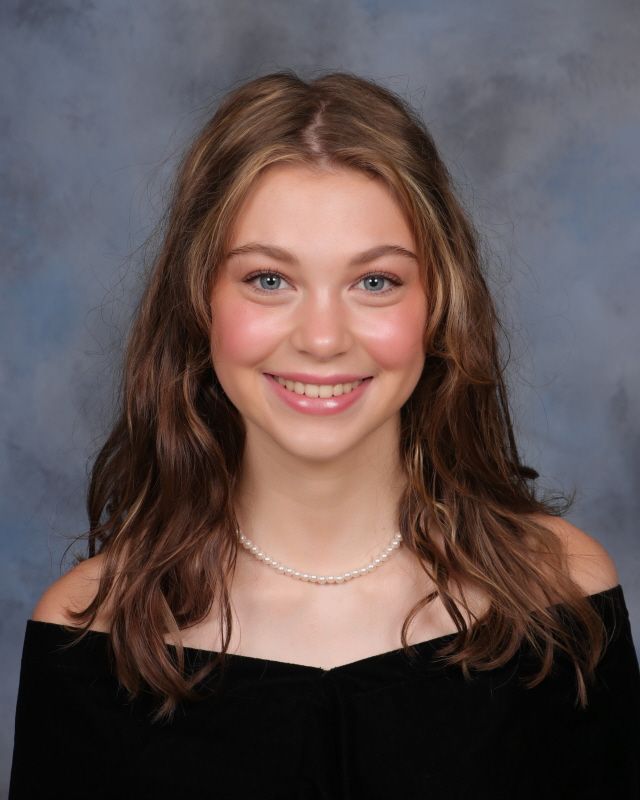 A person with shoulder-length wavy brown hair smiles at the camera, wearing a pearl necklace and a black off-shoulder top.