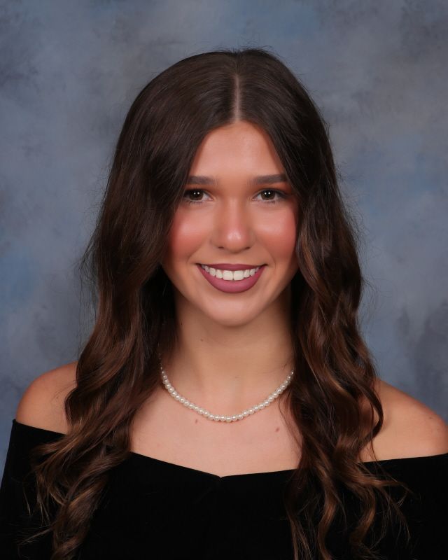 A person with long dark hair, wearing a pearl necklace and black off-the-shoulder top, smiles against a dark background.