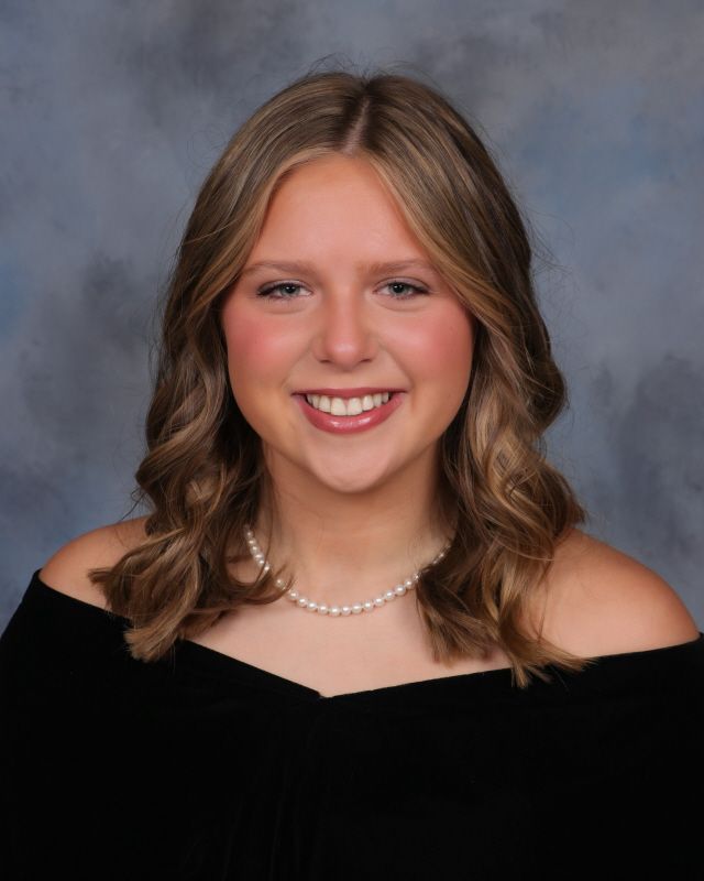 A person with long blonde hair, wearing glasses and a pearl necklace, smiles toward the camera against a brown backdrop.