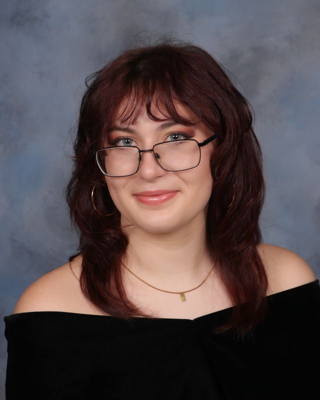 A smiling person with long, dark hair wearing a pearl necklace and a black off-the-shoulder top against a neutral backdrop.