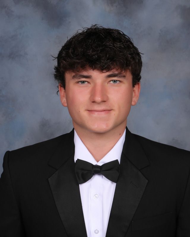 Portrait of a person wearing a grey suit, light shirt, and patterned tie against a plain brown background.
