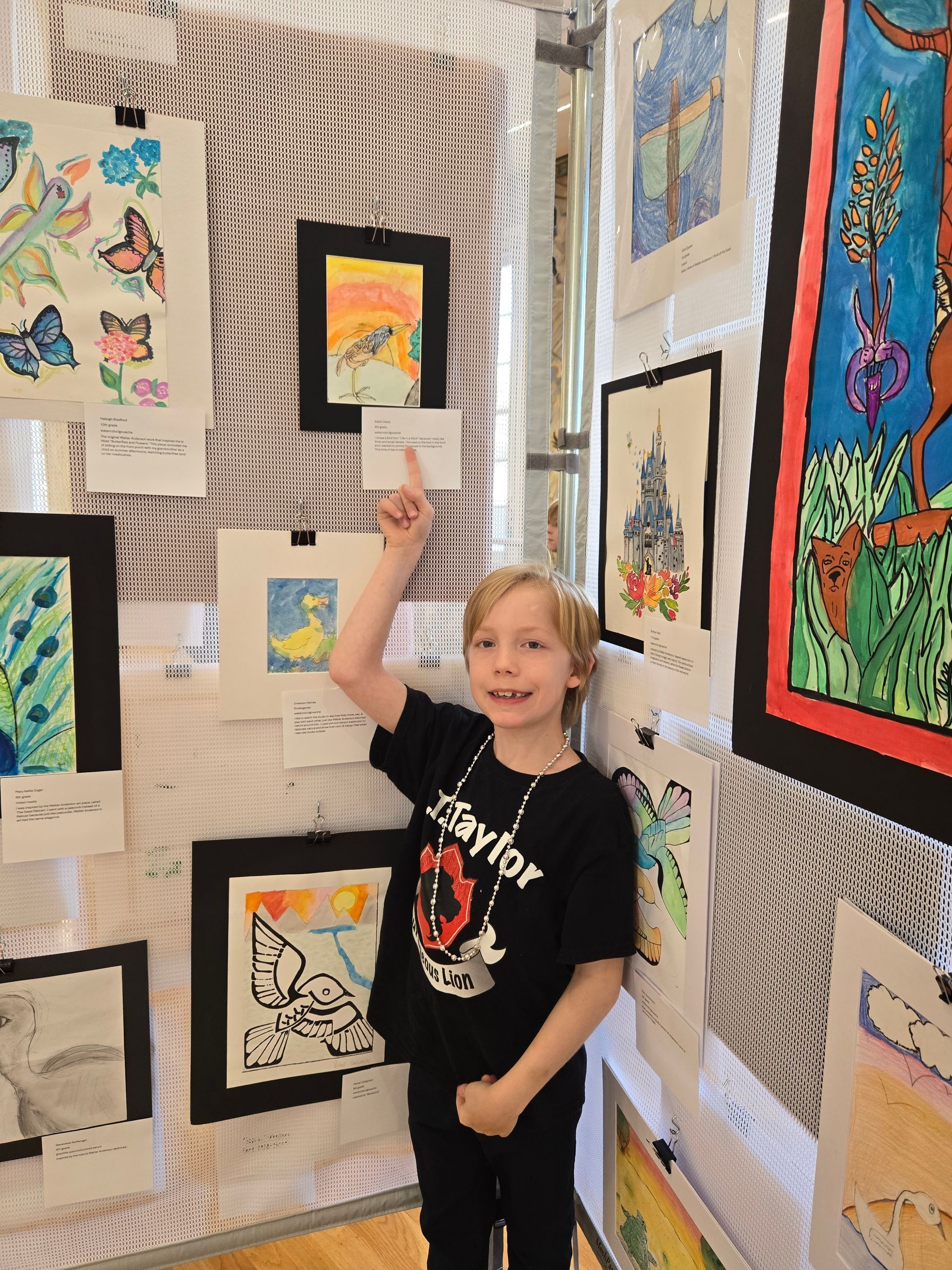 A smiling person in a black t-shirt stands in front of a wall of student art, pointing up at a small framed drawing.