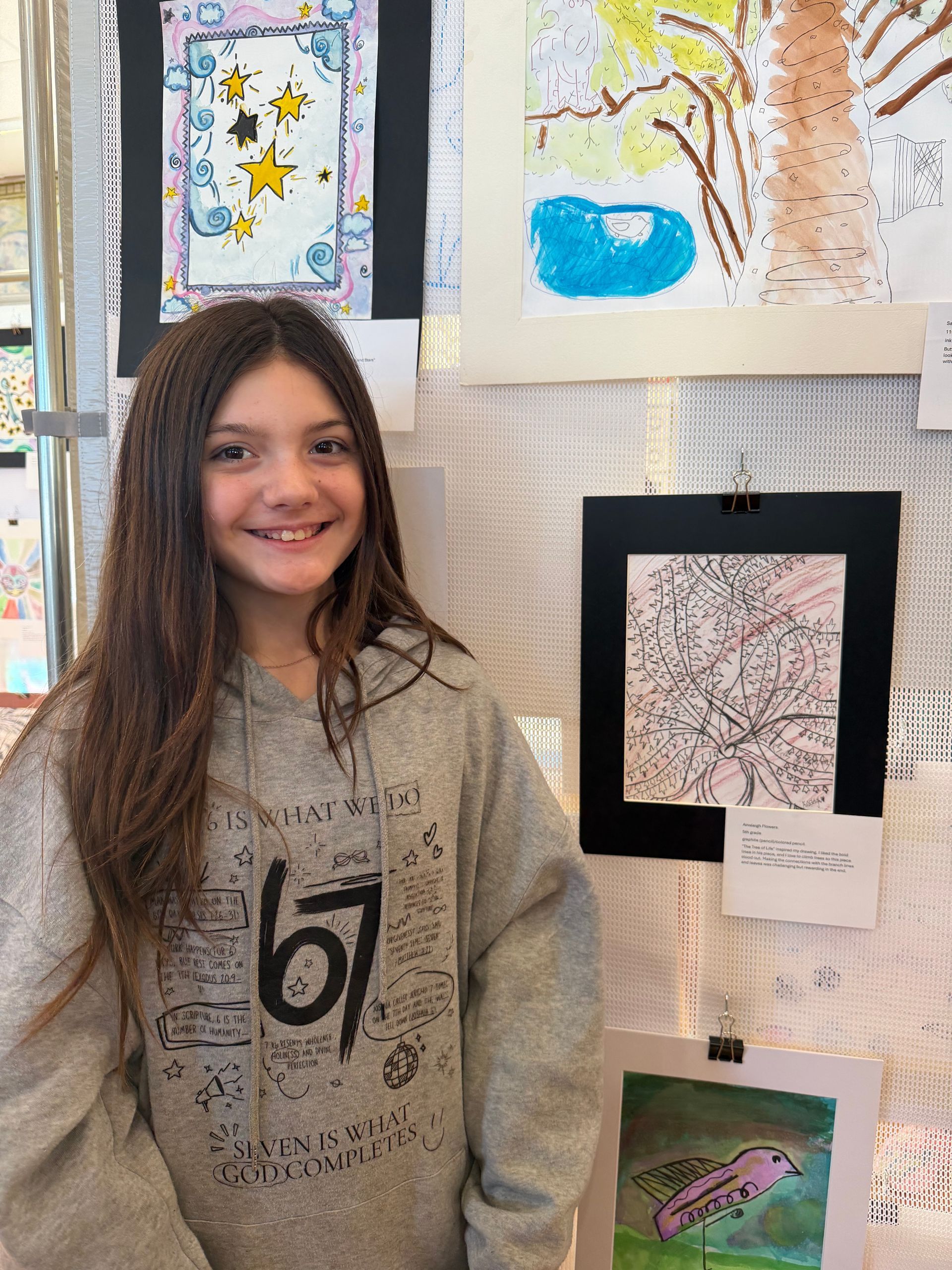 A smiling student stands in an art gallery, posing in front of several framed artworks displayed on a wall.