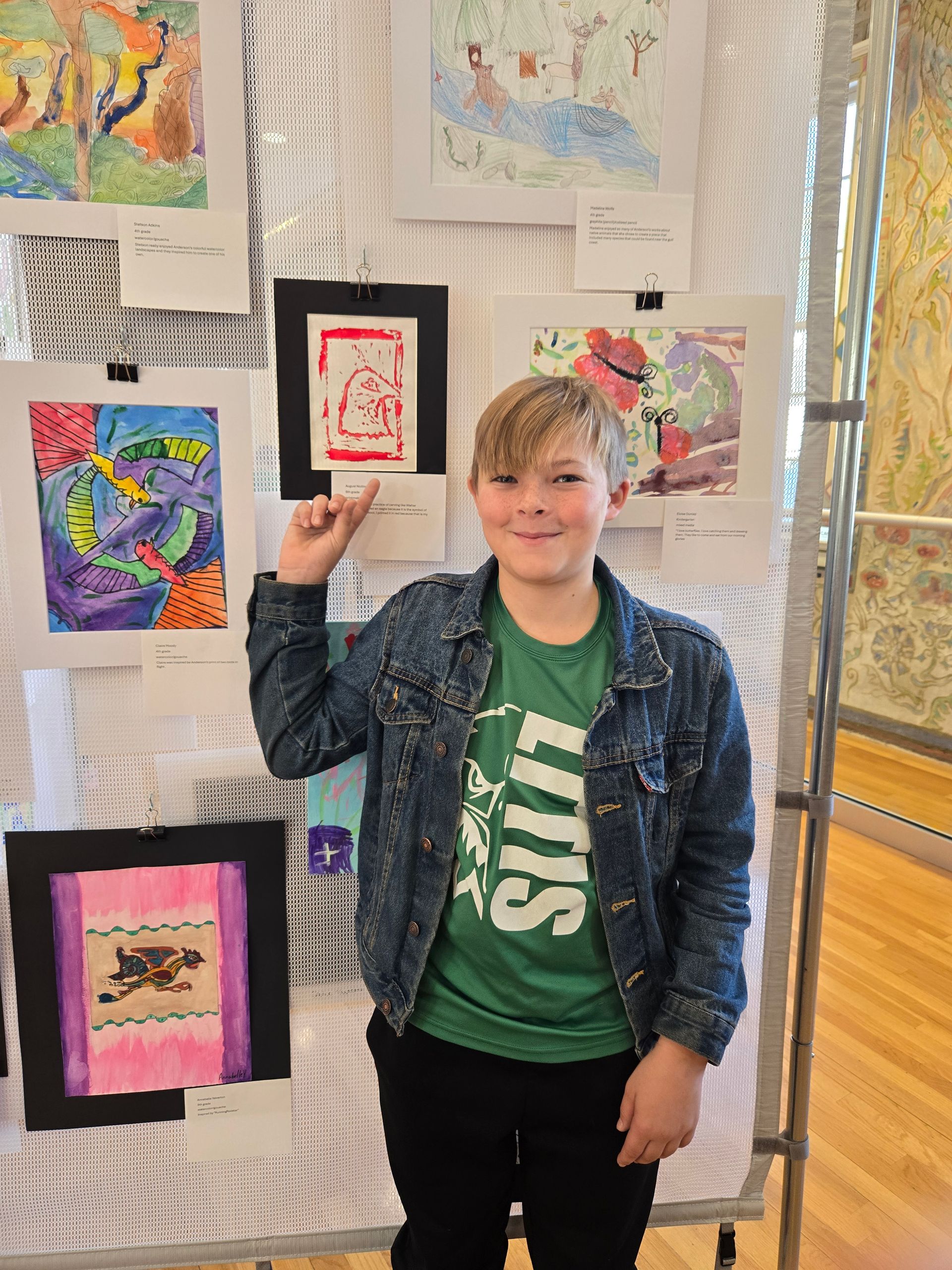 A person stands in front of a gallery wall covered in colorful student artwork, pointing up at a small framed print.