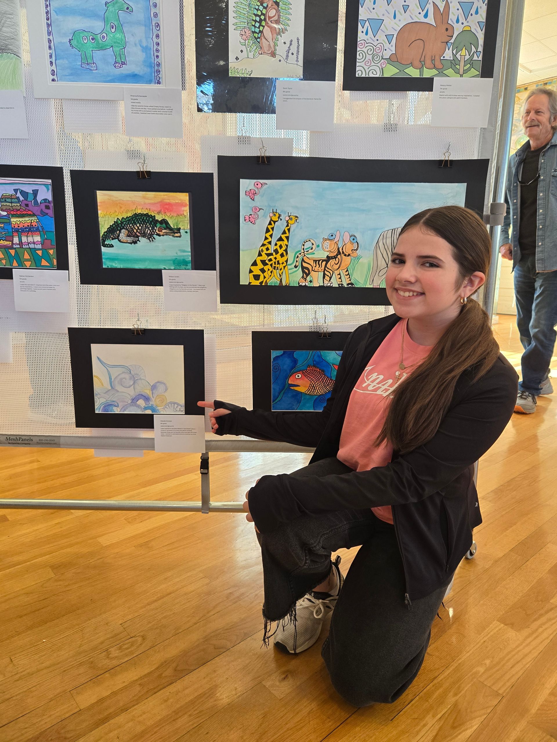 A smiling student kneels in front of an art display featuring several framed paintings on a wire grid in a room.