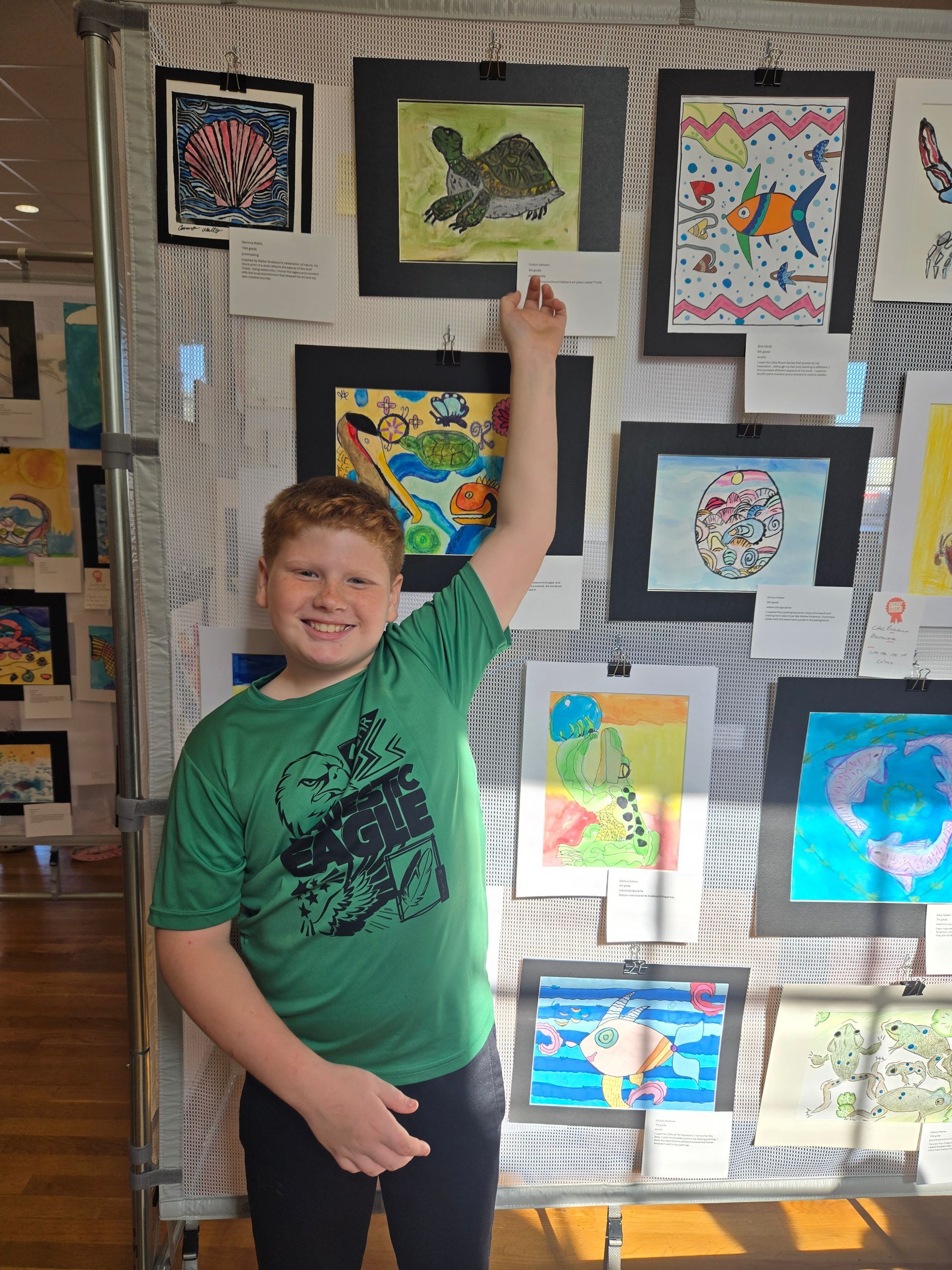 A smiling person wearing a green shirt points to their artwork displayed on a wall with other children's drawings.