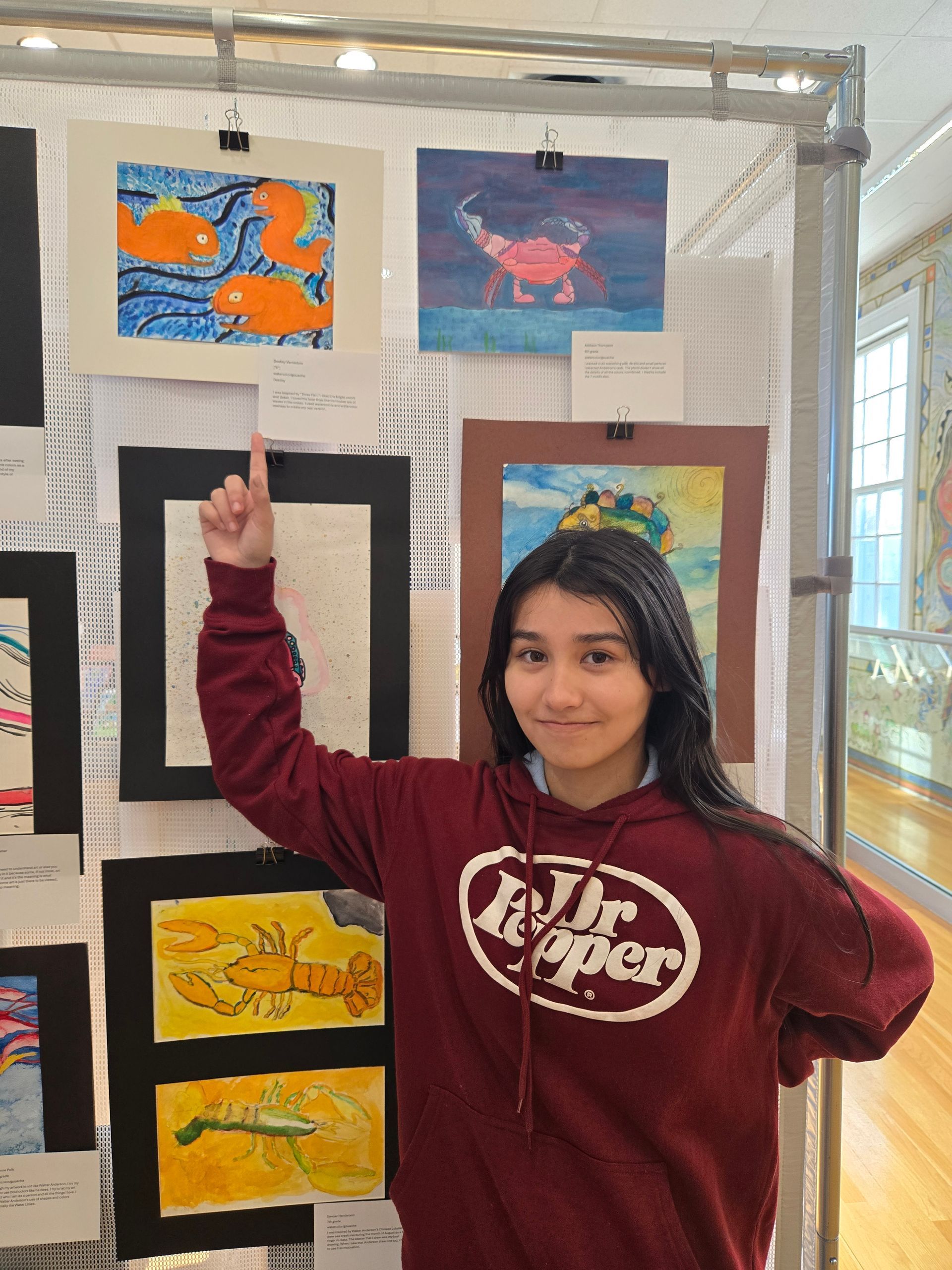 A person in a maroon hoodie points to their artwork displayed on a wire grid panel at an art exhibition.