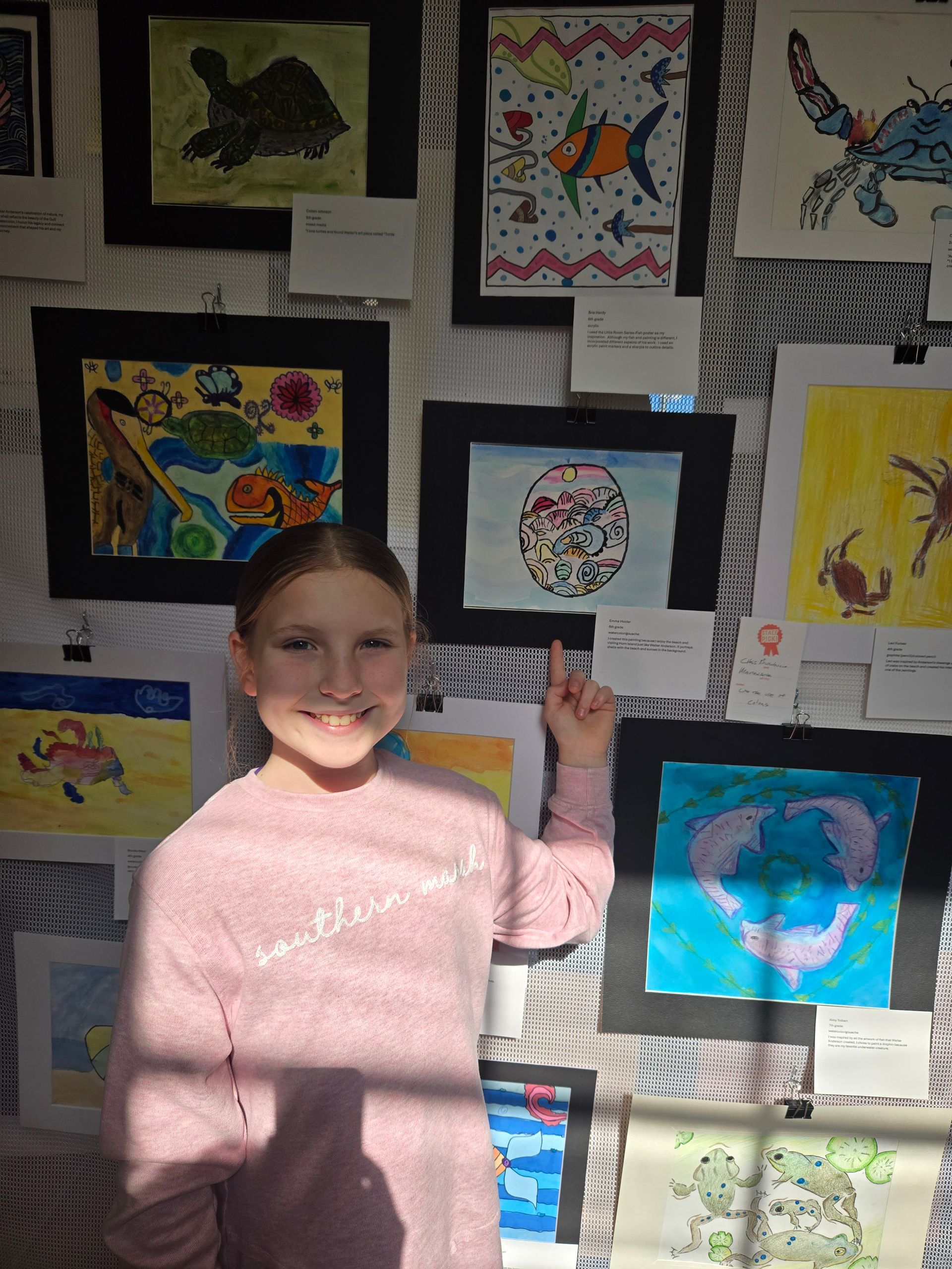 A smiling person stands in front of a wall of colorful children's artwork, pointing to one of the framed drawings.