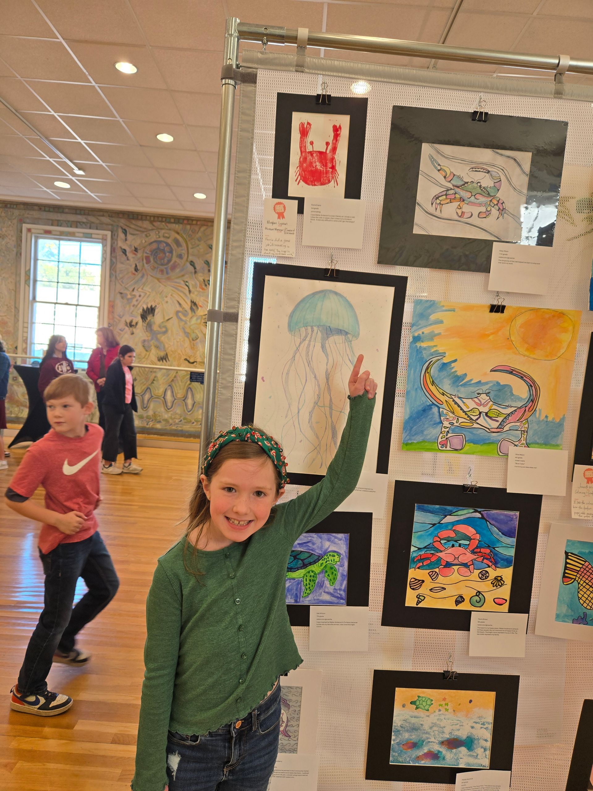 A smiling child points to a drawing of a jellyfish on a display board filled with children's artwork in an indoor room.
