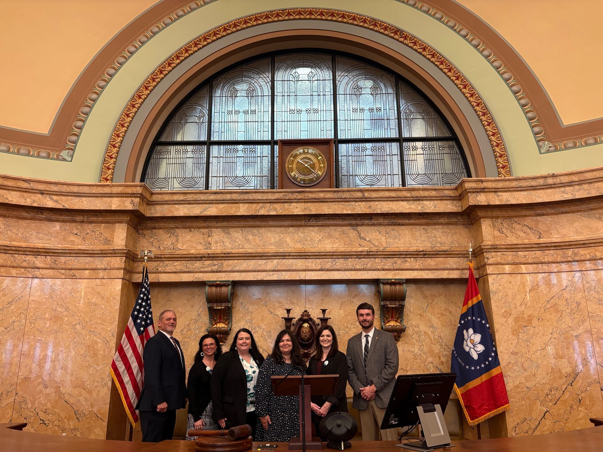 A group of six people stands behind a wooden podium inside a legislative chamber, flanked by a U.S. and Mississippi flag.