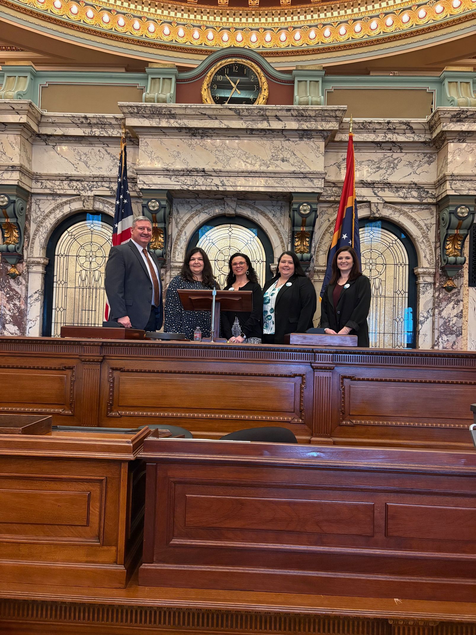 Five people stand behind a wooden podium in a grand, marble legislative chamber with U.S. and state flags.