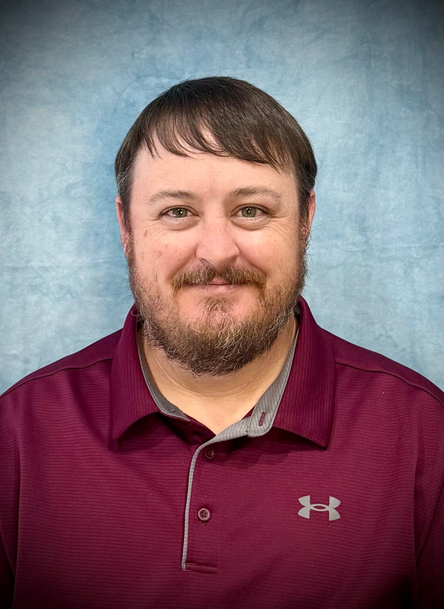A person with short brown hair and a beard, wearing a maroon polo shirt against a light blue, textured backdrop.