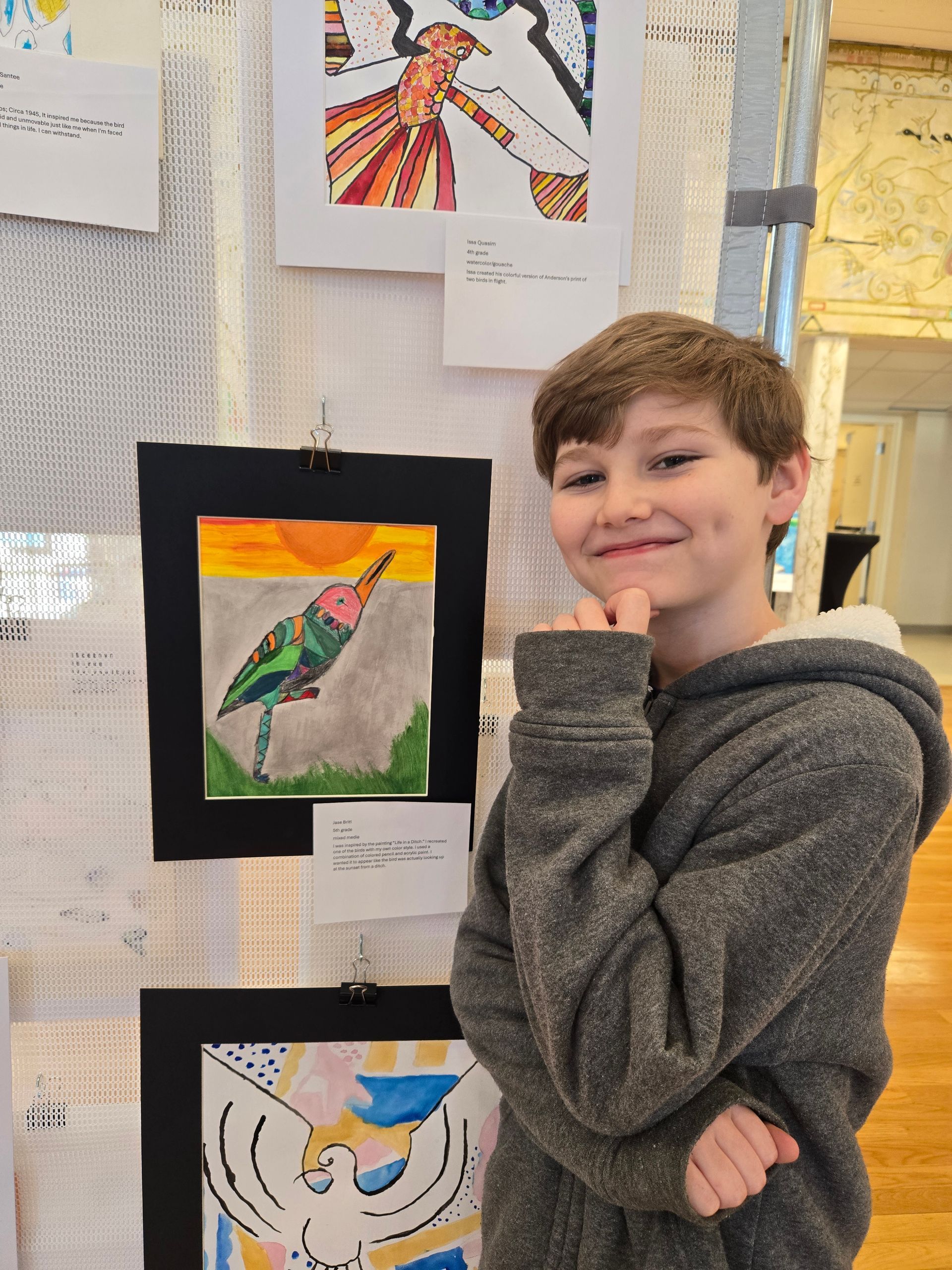 A person smiling and posing next to their colorful artwork displayed on a wall.