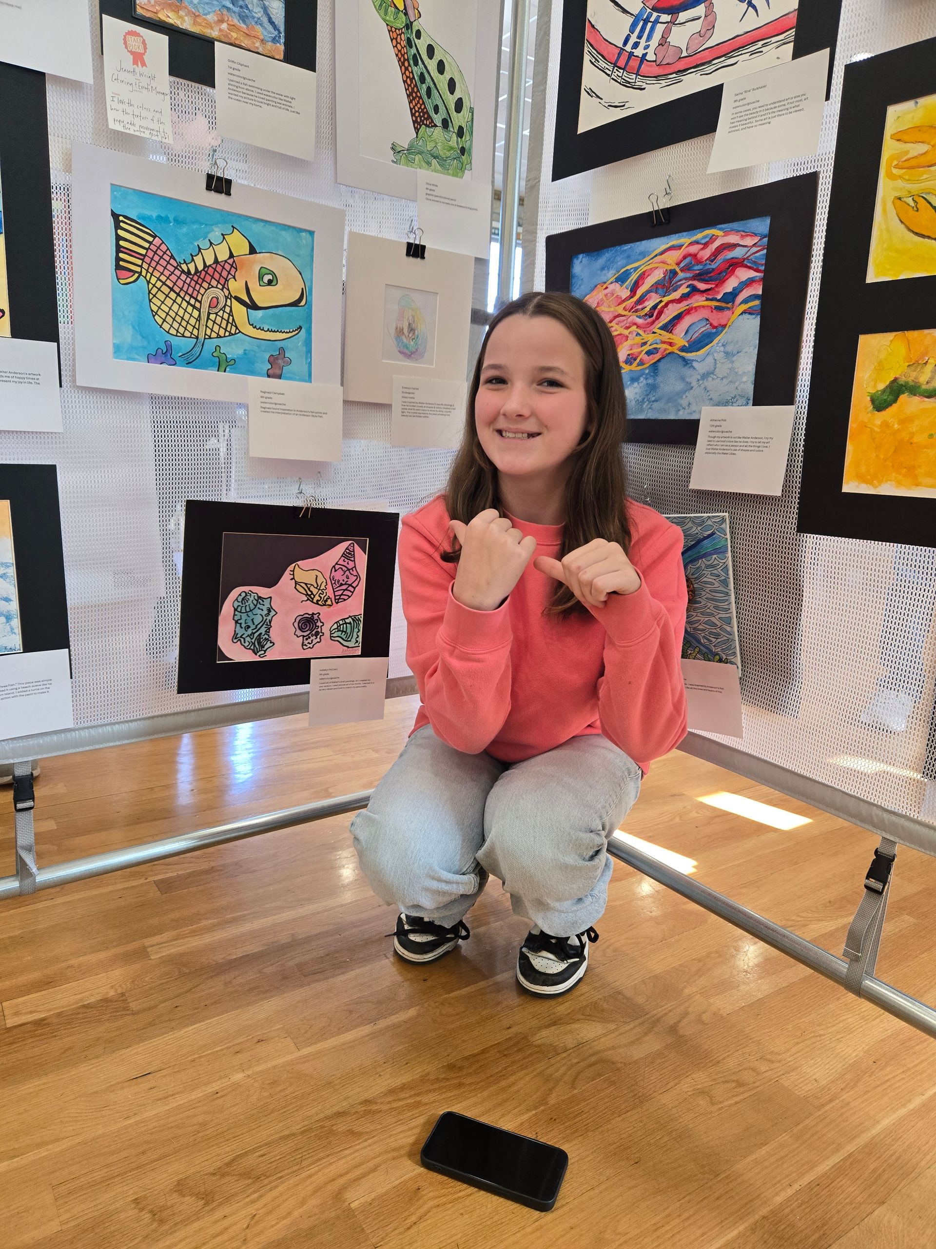 A smiling person crouches in front of a display of framed student artwork, gesturing toward a piece behind them.