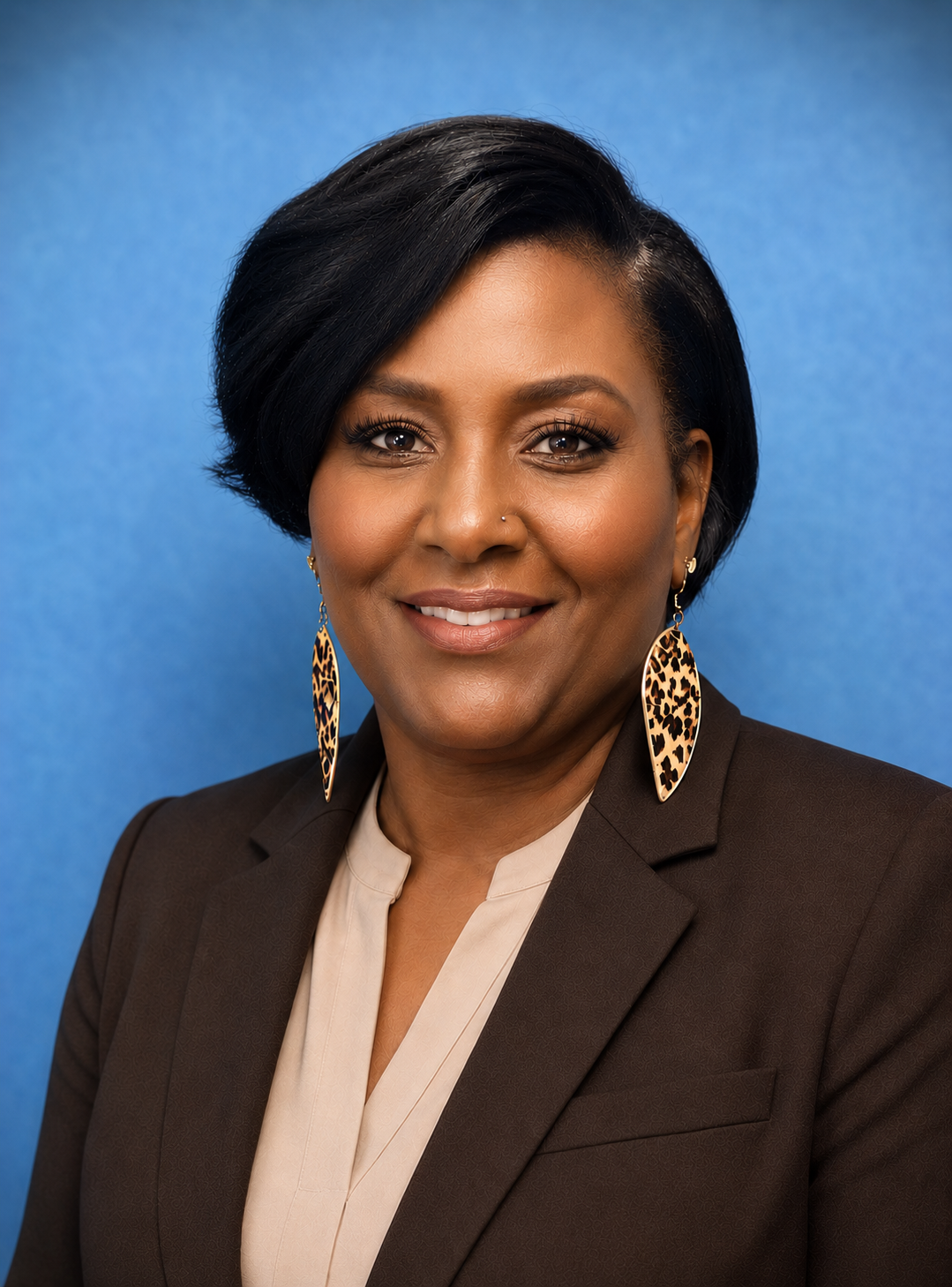 A person with dark hair and leopard print earrings, wearing a brown top and tan cardigan against a blue textured backdrop.
