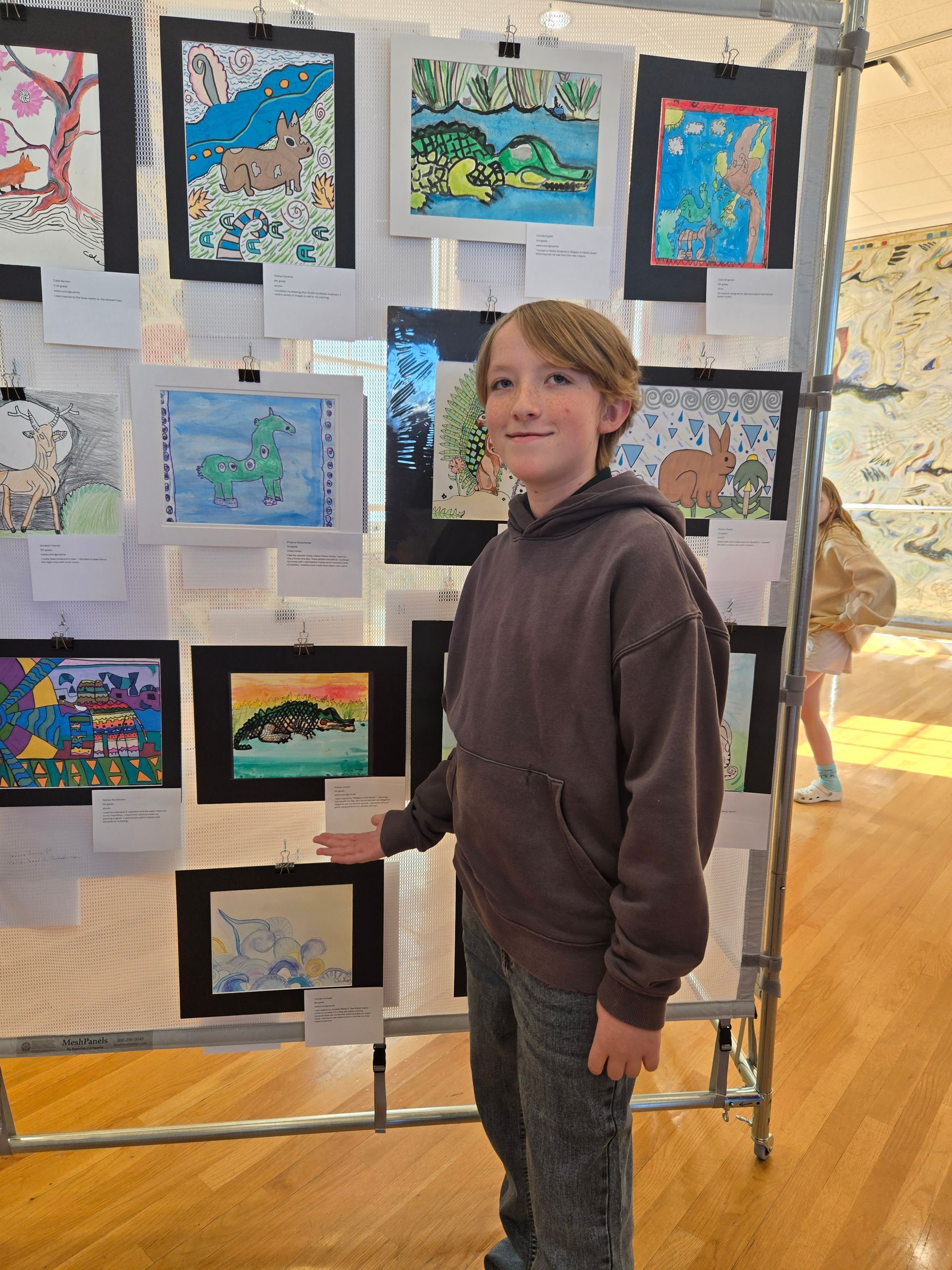 A youth in a brown hoodie stands in front of a grid display of colorful drawings pinned to a frame at an art exhibition.