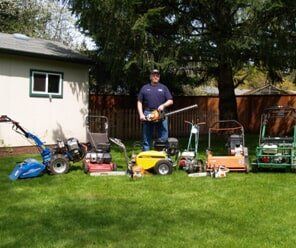 Man stands with lawn equipment in a yard. Tools include a tiller, edger, and mower.
