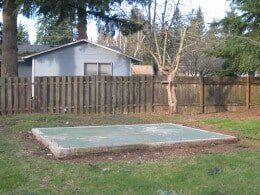 A concrete pad in a grassy yard, with a wooden fence and a light blue building in the background.