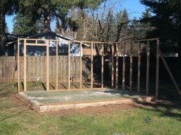 Wooden frame of a building under construction on a concrete base in a backyard.
