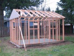 Wooden shed under construction in a yard, with visible framing and roof rafters.