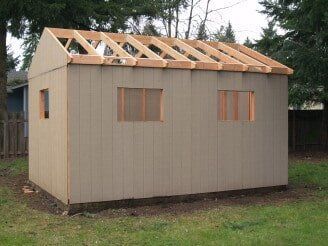 A tan shed under construction with an unfinished wooden roof frame.