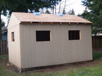 Tan shed with three window openings and a partially built roof in a grassy yard.