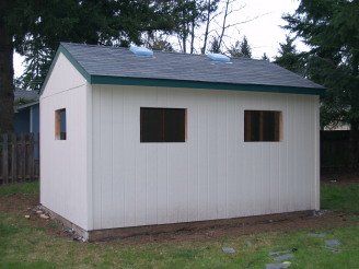 White shed with three windows, green trim, and a gray roof in a grassy backyard.