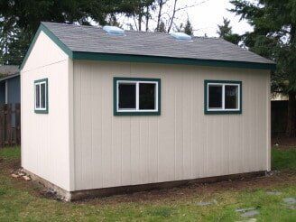 Beige shed with three windows and a gray roof, in a grassy backyard.