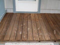 Wooden porch with brown boards in front of a white window with blue trim.