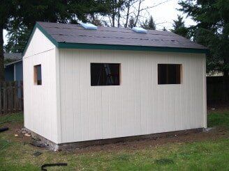 White shed with green trim, dark roof, and three window openings sits on grass.