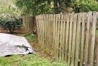 Wooden picket fence in a grassy yard, with a tarp-covered object and trees visible in the background.