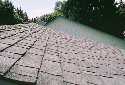 Close-up of a damaged asphalt shingle roof, with raised and uneven shingles.