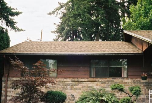 Brown house with brick base and brown roof, two windows, and lush green trees in the background.